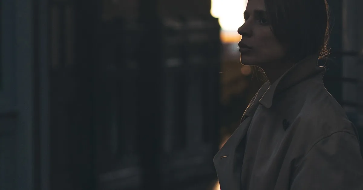 Mulher de perfil, com expressão pensativa, olhando em direção a uma luz quente de fim de tarde, em ambiente escuro e introspectivo