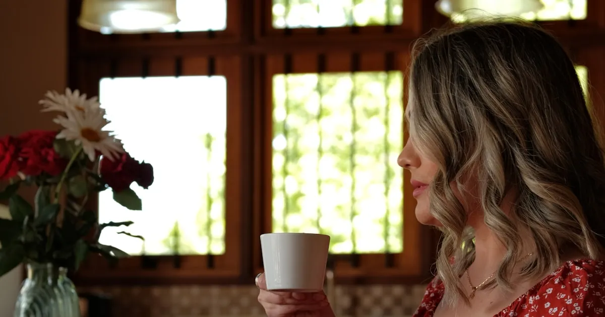 Mulher em uma pausa de silêncio em casa, com uma xícara de café nas mãos, diante da janela iluminada pela luz natural da manhã