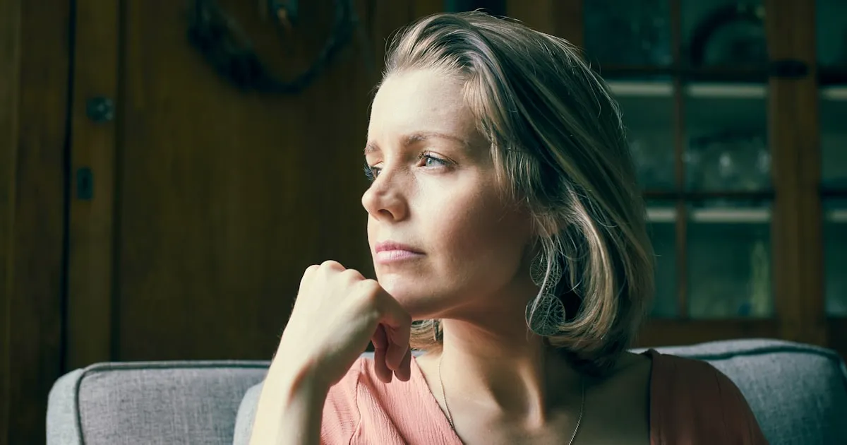 Mulher entre 35 e 55 anos sentada numa poltrona clara, com a mão apoiada perto do queixo e o olhar voltado para a janela, num momento de pausa e reflexão sob luz natural