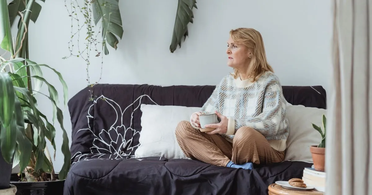 Mulher na faixa dos 50 anos sentada sozinha no sofá de casa, com uma xícara nas mãos e o olhar voltado para longe, em luz natural suave