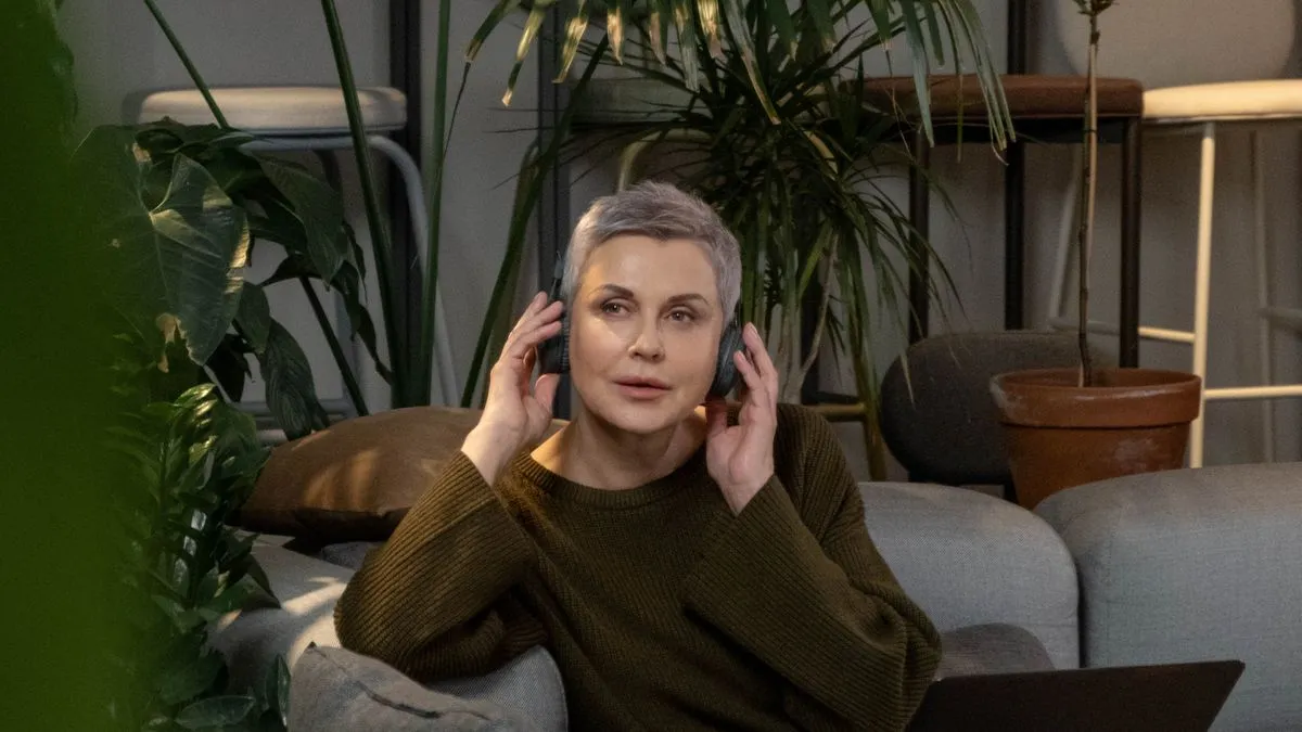 Mulher madura sentada no sofá de casa, ajustando o fone de ouvido diante do notebook, cercada de plantas, no começo de uma sessão de terapia online.