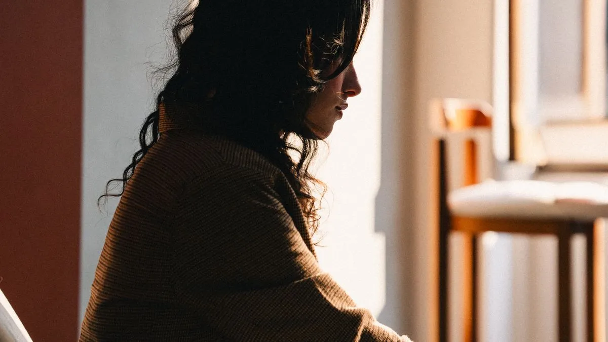 Mulher de perfil sentada num canto silencioso da casa, banhada pela luz quente do fim da tarde, num momento de pausa em que finalmente escuta o que está sentindo