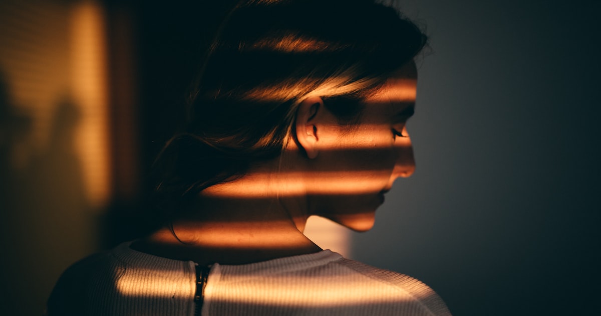 Mulher em perfil, de olhos baixos, banhada pela luz quente e listrada de uma persiana, num momento de introspecção