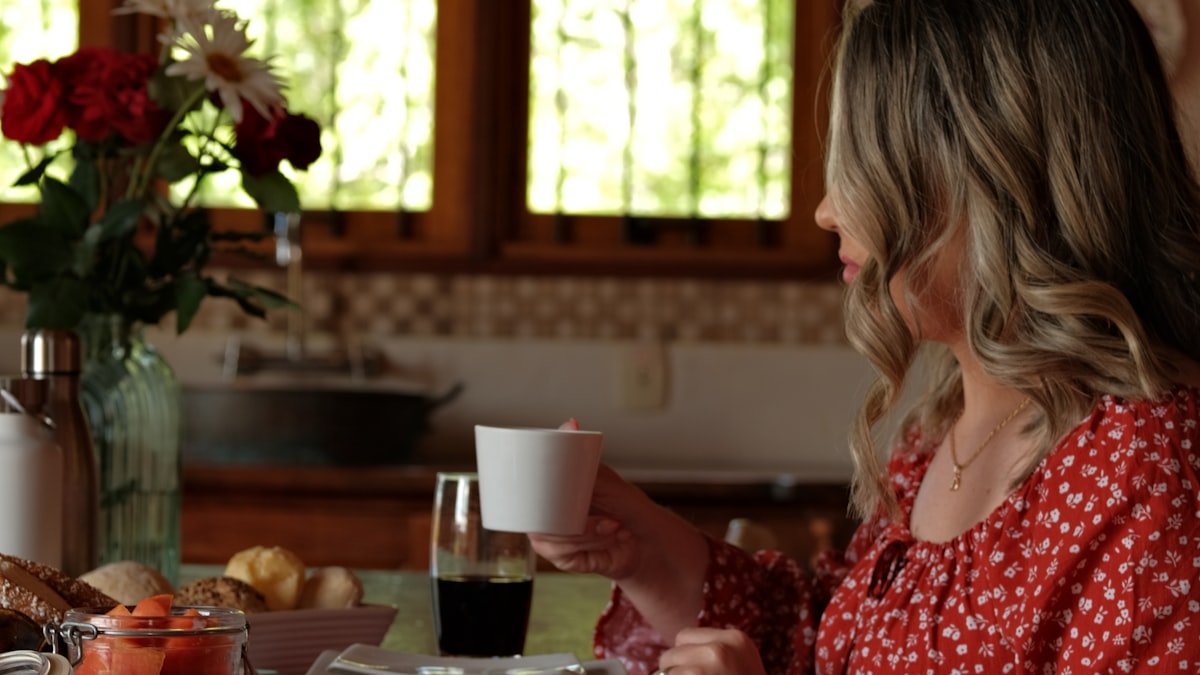 Mulher sentada à mesa posta da própria casa, com uma xícara nas mãos e a luz da manhã entrando pela janela, num café tomado sentada e sem pressa