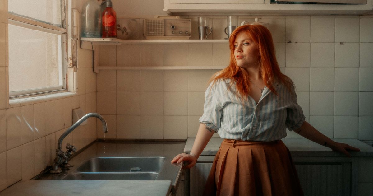Mulher parada na cozinha de casa, ao lado da pia e da janela por onde entra a luz quente da tarde, com o olhar voltado para longe num momento de pausa
