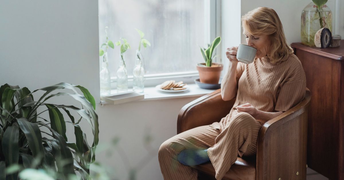 Mulher sentada sozinha na poltrona perto da janela, tomando um café devagar, em uma casa silenciosa com luz natural suave.