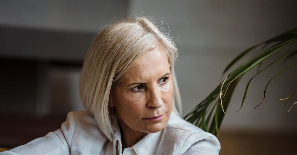Mulher na faixa dos 50 anos sentada no sofá de casa, de camisa branca, com o olhar voltado para o lado e a expressão contida de quem está pensando em algo que ainda não disse em voz alta