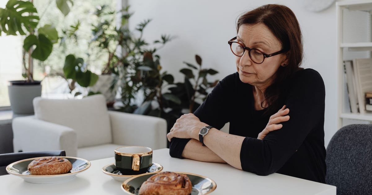 Mulher de meia-idade sentada sozinha à mesa de casa, de braços cruzados sobre o tampo, com o café e o pão servidos e intocados na frente dela, olhando para baixo em silêncio.