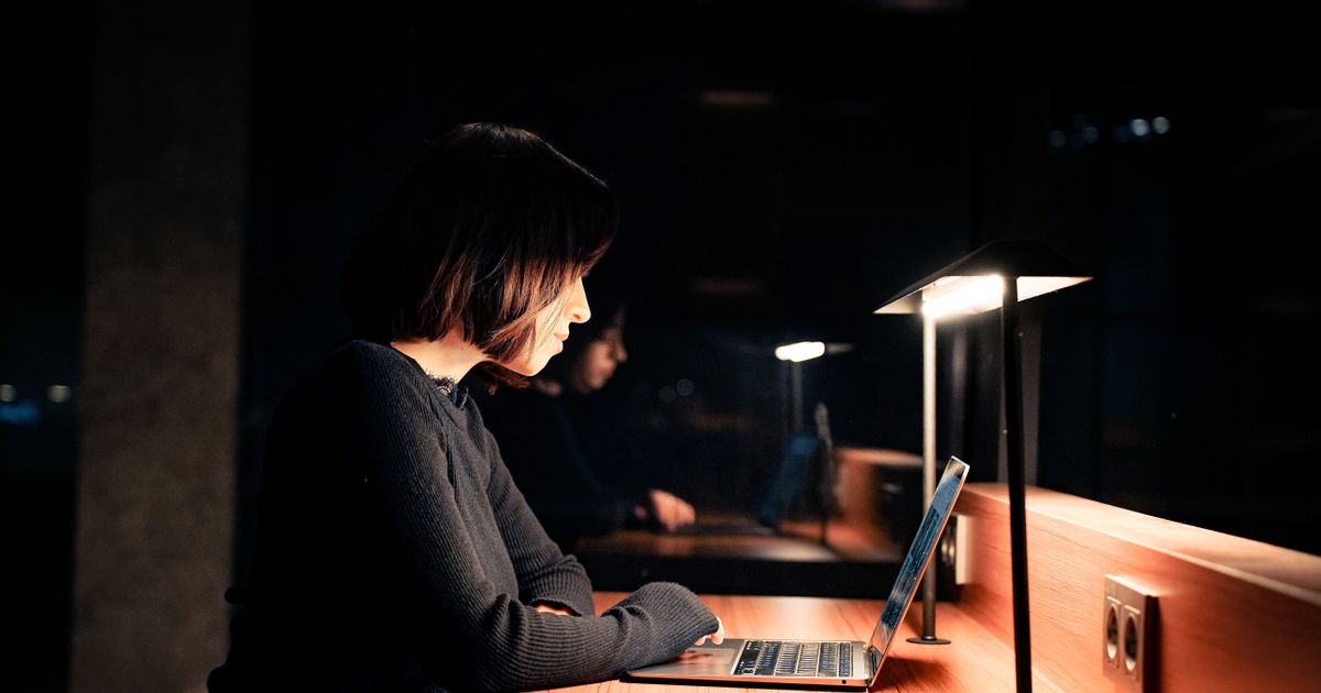 Mulher trabalhando sozinha à noite, iluminada apenas pela luminária da mesa e pela tela do notebook, com o olhar cansado de quem continua mesmo depois do expediente