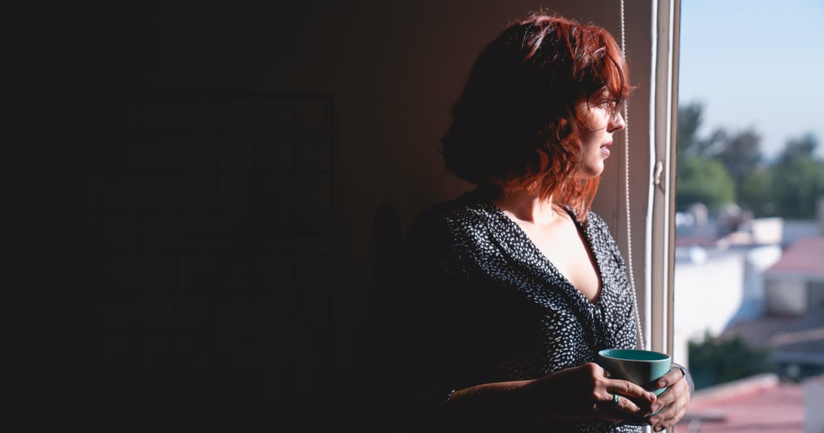 Mulher de pé junto à janela, com uma caneca nas mãos e o olhar perdido lá fora, banhada pela luz quente da manhã