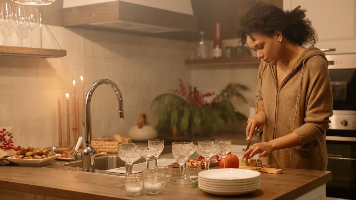 Mulher sozinha na cozinha, sob luz quente, preparando a mesa e a comida para os outros enquanto ninguém aparece por perto