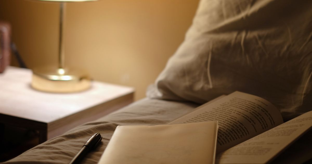 Abajur aceso na mesa de cabeceira ao lado da cama, com livro aberto e caderno sobre o edredom, à noite, sem nenhum celular por perto.