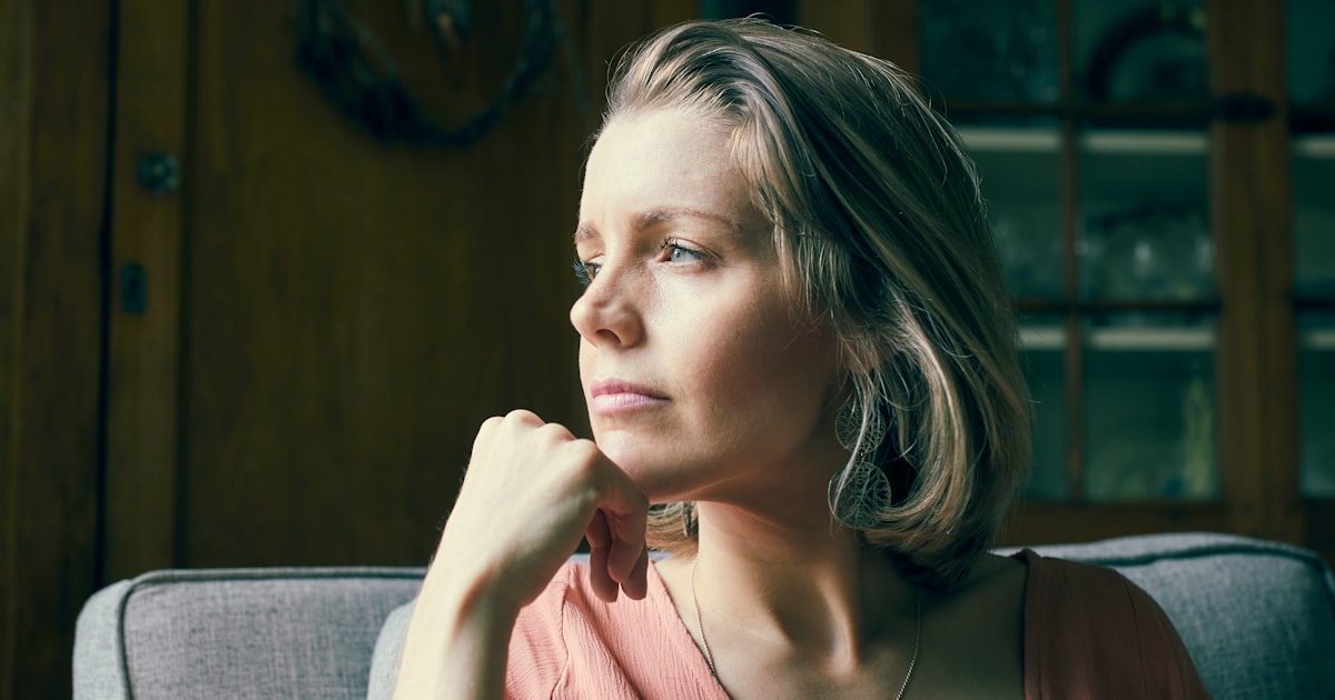 Mulher entre 35 e 55 anos sentada numa poltrona clara, com a mão apoiada perto do queixo e o olhar voltado para a janela, num momento de pausa e reflexão sob luz natural