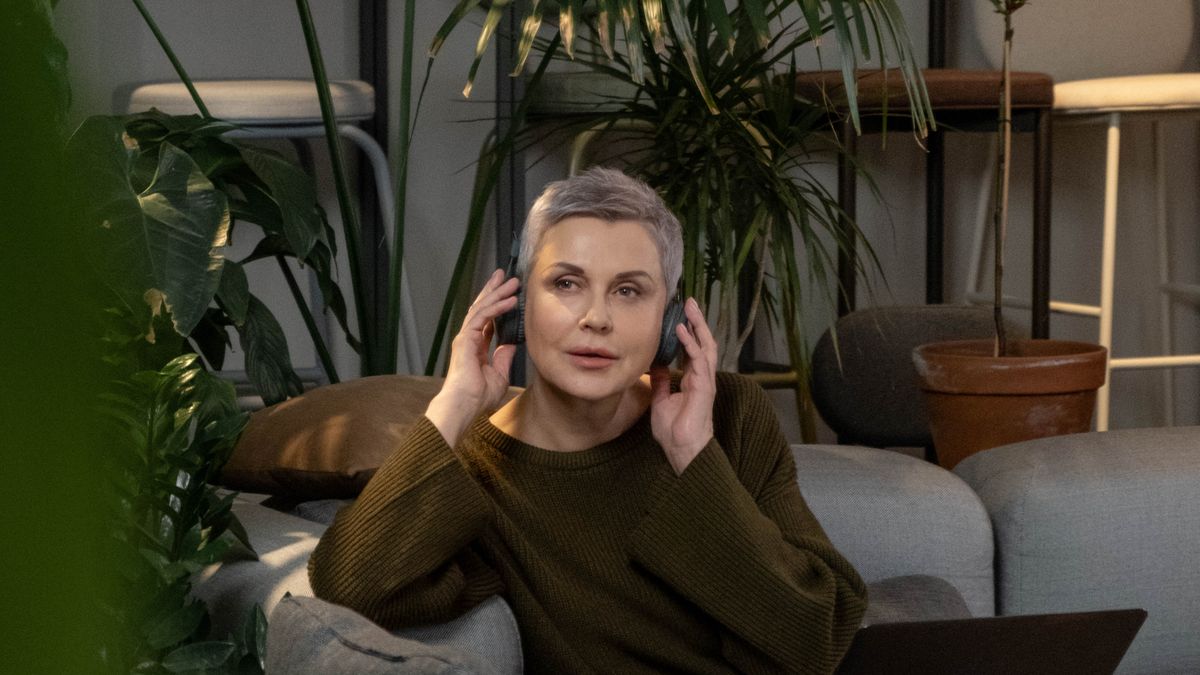 Mulher madura sentada no sofá de casa, ajustando o fone de ouvido diante do notebook, cercada de plantas, no começo de uma sessão de terapia online.