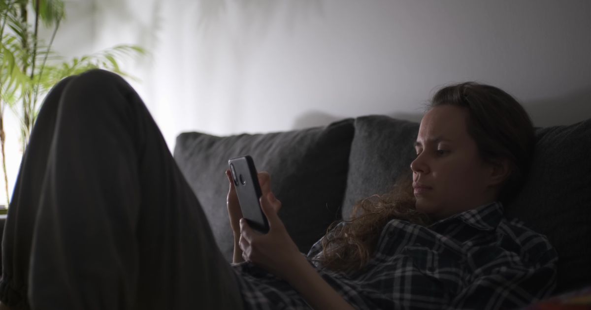 Mulher sentada no sofá da sala à noite, com o celular na mão e a luz baixa do abajur, olhando para a tela com expressão de hesitação antes de enviar uma mensagem