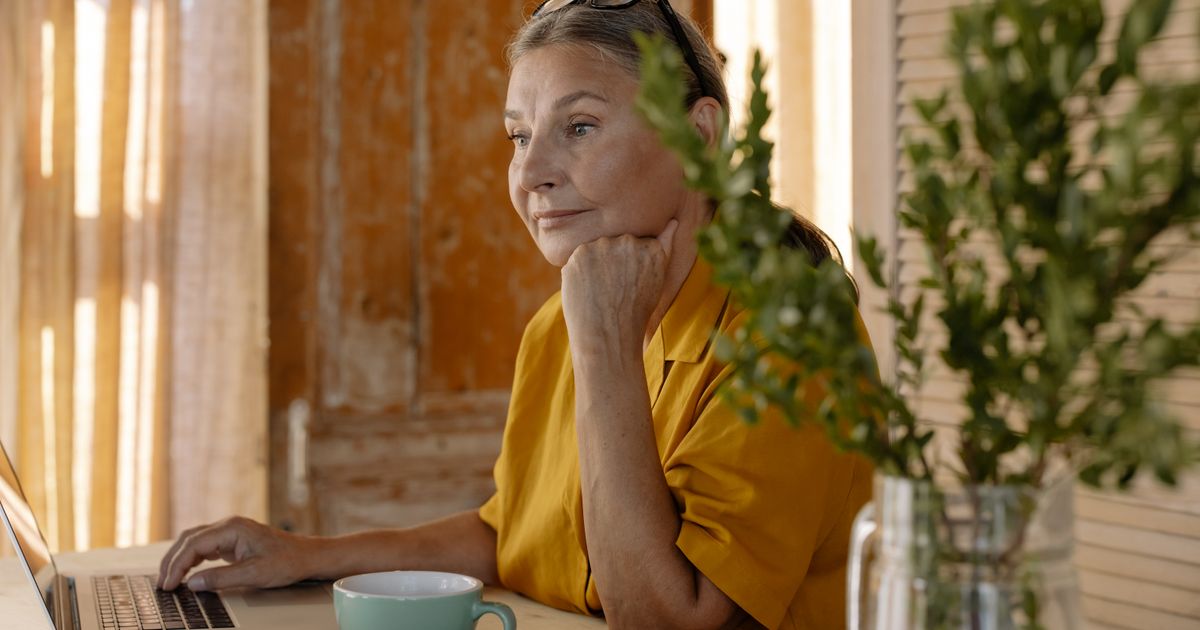 Mulher de meia-idade sentada à mesa de casa, com a mão apoiada no queixo e o olhar pensativo, banhada pela luz natural quente da manhã