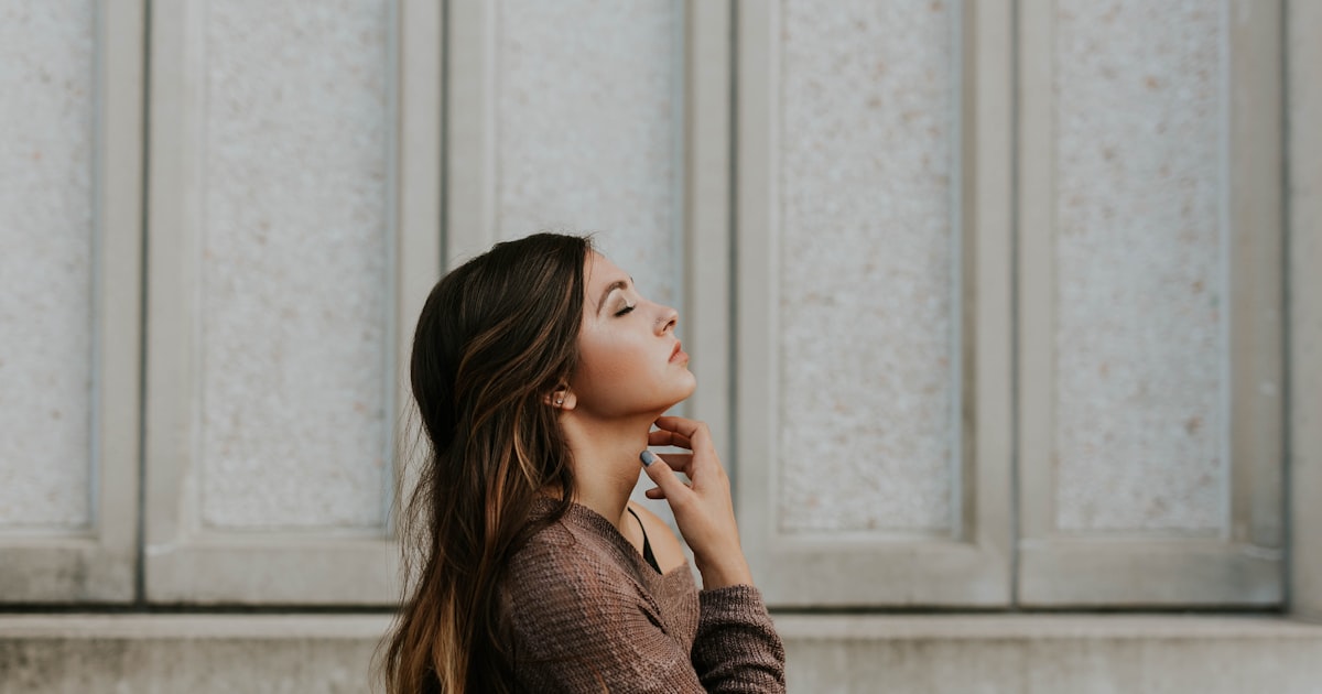 Mulher de perfil junto a uma parede clara, em luz natural suave, olhos fechados e mão no rosto, num momento de respiro e introspecção