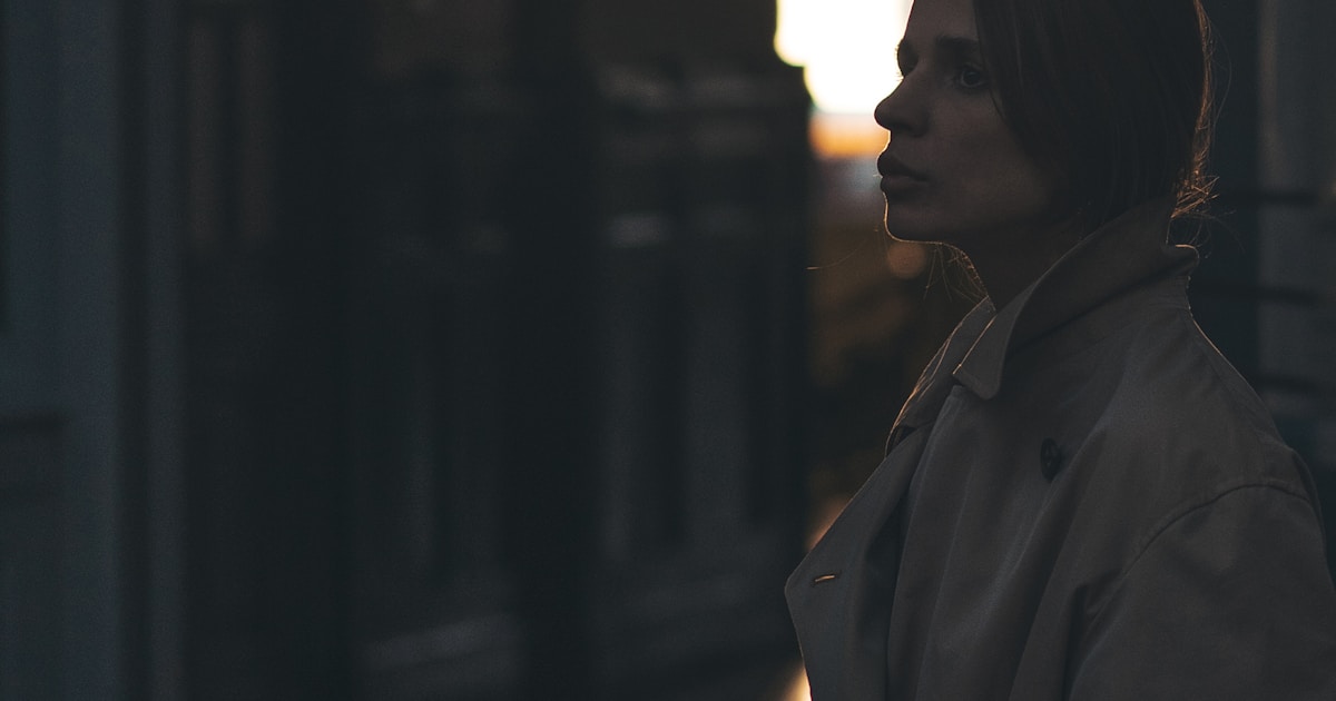 Mulher de perfil, com expressão pensativa, olhando em direção a uma luz quente de fim de tarde, em ambiente escuro e introspectivo