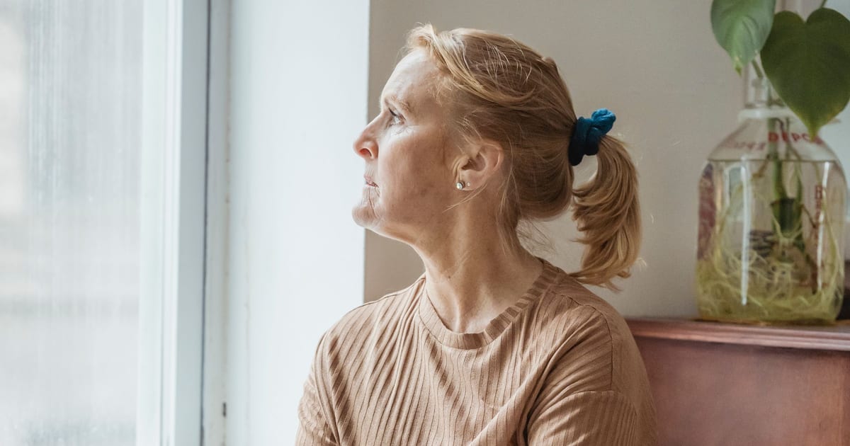 Mulher na faixa dos 45 anos sentada em uma poltrona junto à janela, de perfil, em luz natural suave, com o olhar voltado para fora num momento de reflexão