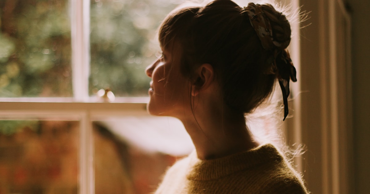 Mulher de perfil olhando pela janela, sob luz natural quente, num momento de reflexão