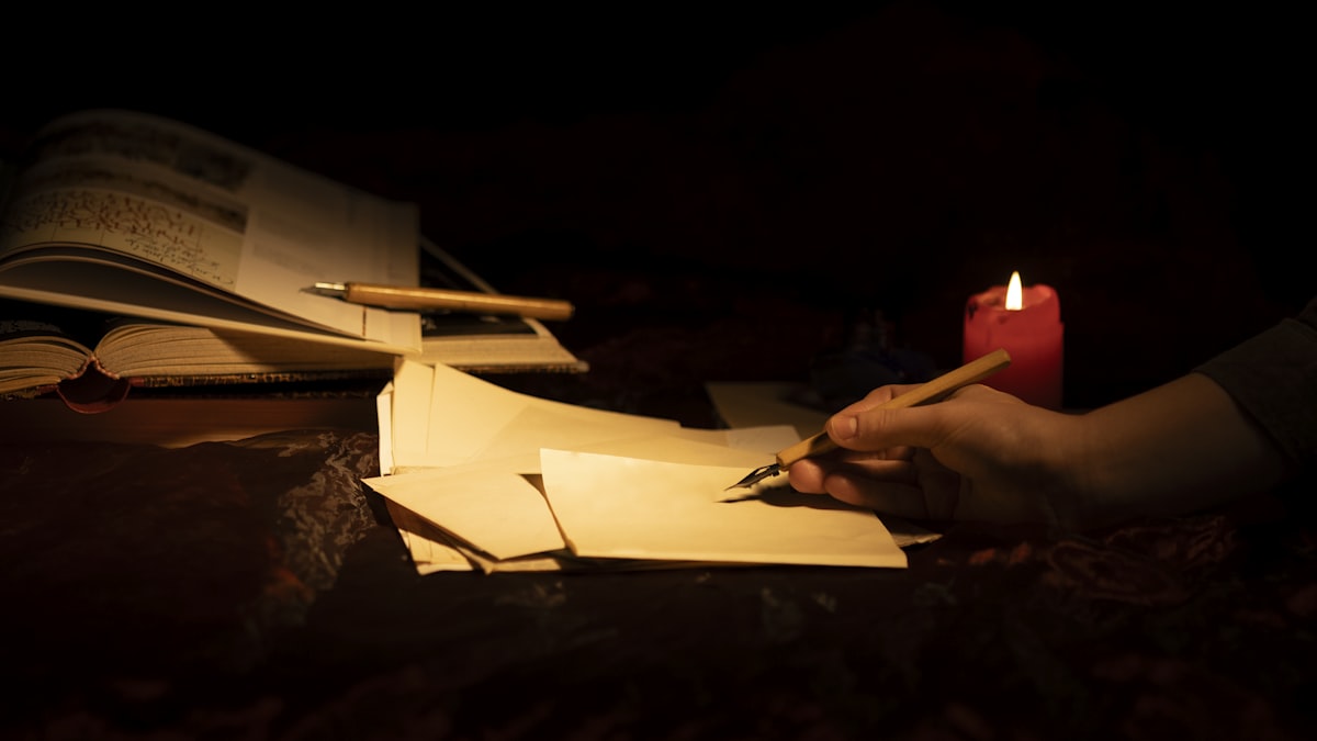 Mão escrevendo à luz de vela sobre papel, com um caderno aberto ao lado, num gesto íntimo de escuta de si.