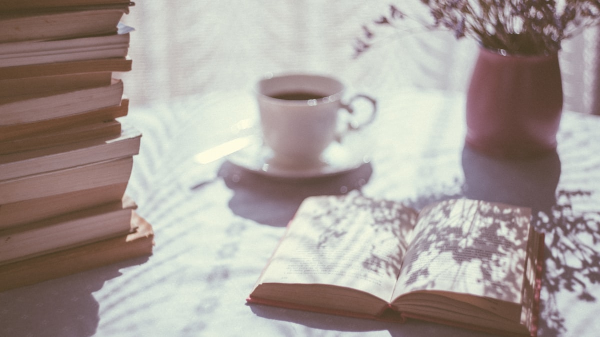 Livro aberto ao lado de uma xícara de café e flores, sob luz suave da manhã, num clima calmo de leitura sem pressa