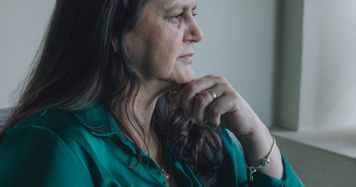 Mulher por volta dos 50 anos sentada perto da janela de casa, de perfil, com a mão apoiada no queixo e o olhar parado, no silêncio de quem está tentando entender o que sente