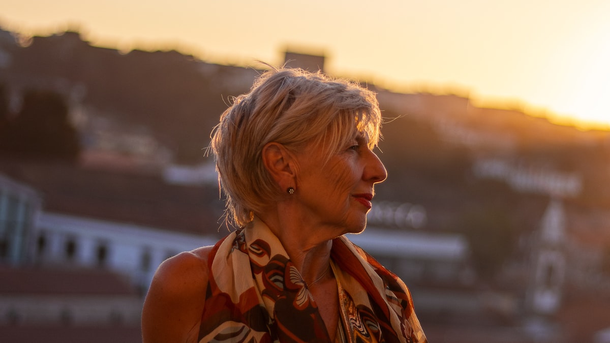 Mulher por volta dos 55 anos ao ar livre, de perfil, com o olhar voltado para longe sob a luz dourada do fim de tarde, num momento de reencontro consigo