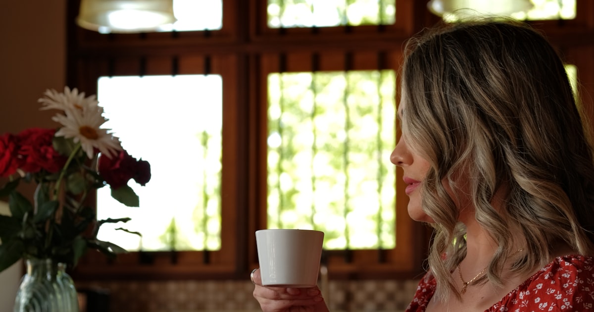 Mulher em uma pausa de silêncio em casa, com uma xícara de café nas mãos, diante da janela iluminada pela luz natural da manhã