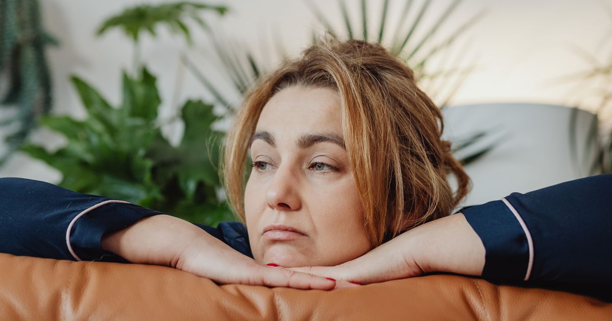 Mulher por volta dos quarenta anos apoiada no encosto do sofá de couro caramelo, com o queixo sobre as mãos e o olhar distante, em um momento de pausa dentro de casa