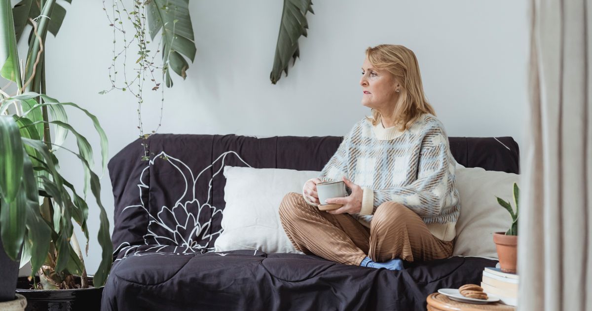 Mulher na faixa dos 50 anos sentada sozinha no sofá de casa, com uma xícara nas mãos e o olhar voltado para longe, em luz natural suave