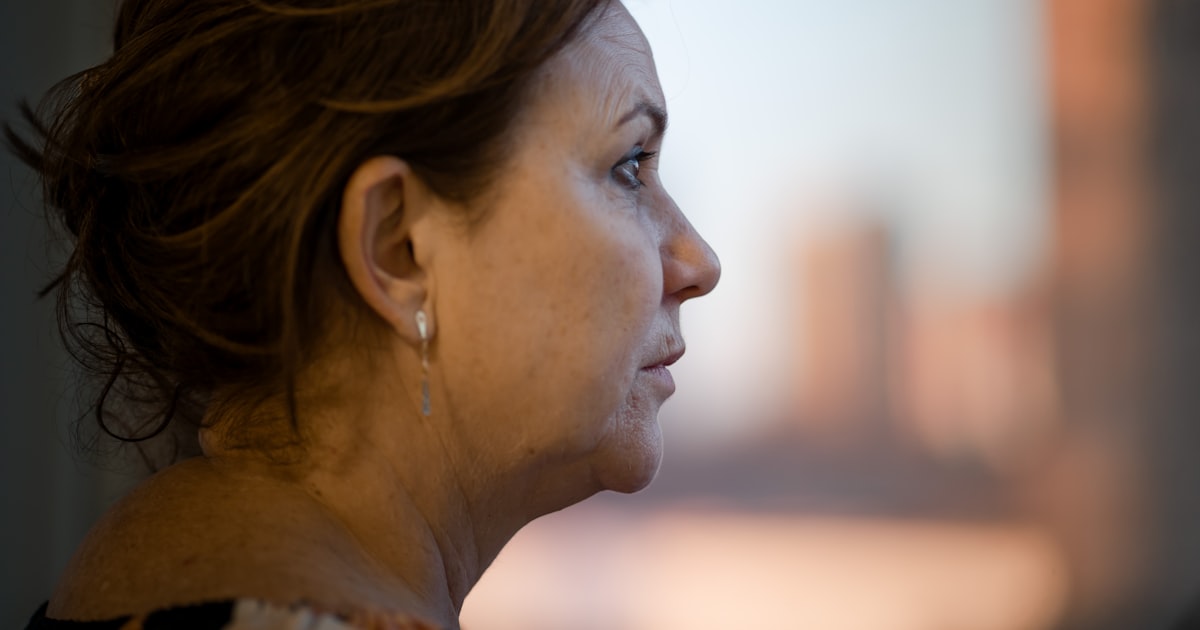 Mulher por volta dos 50 anos, de perfil junto a uma janela ao entardecer, com olhar pensativo e sereno, em luz natural quente