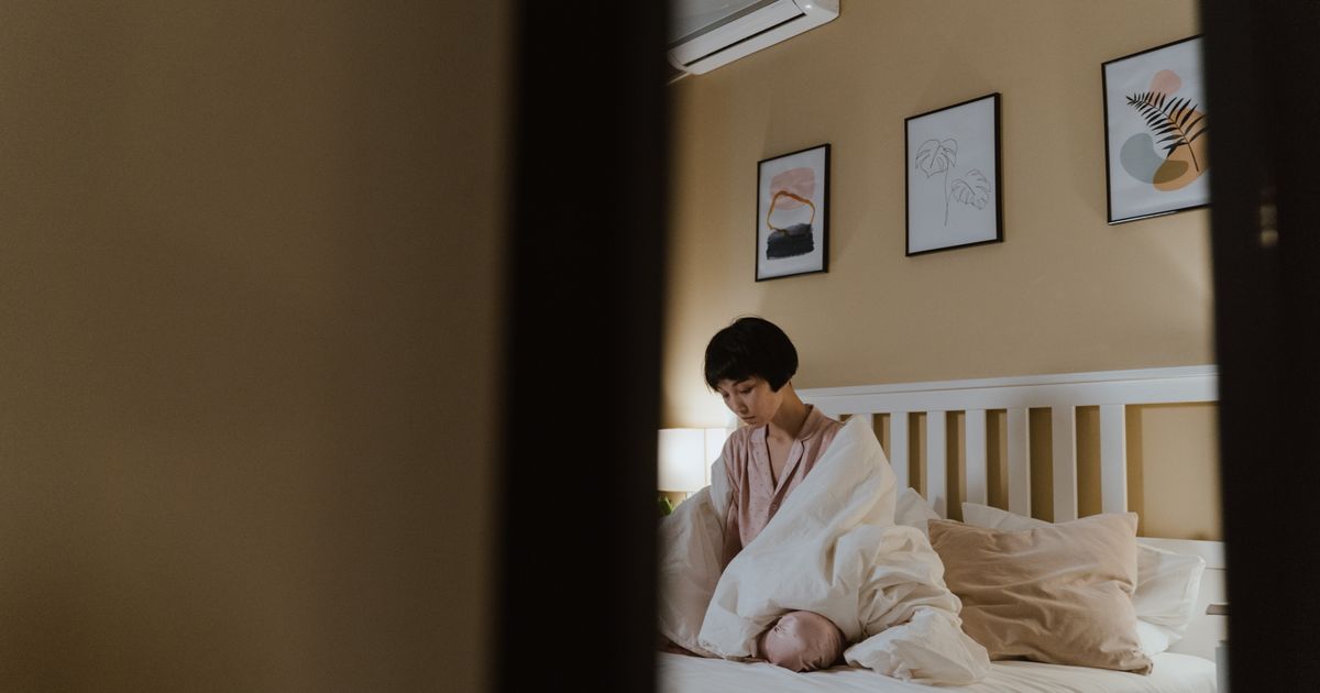 Mulher sentada sozinha na beira da cama de um quarto arrumado, com o olhar baixo e distante, na luz quente do abajur no fim do dia, vista através do vão da porta